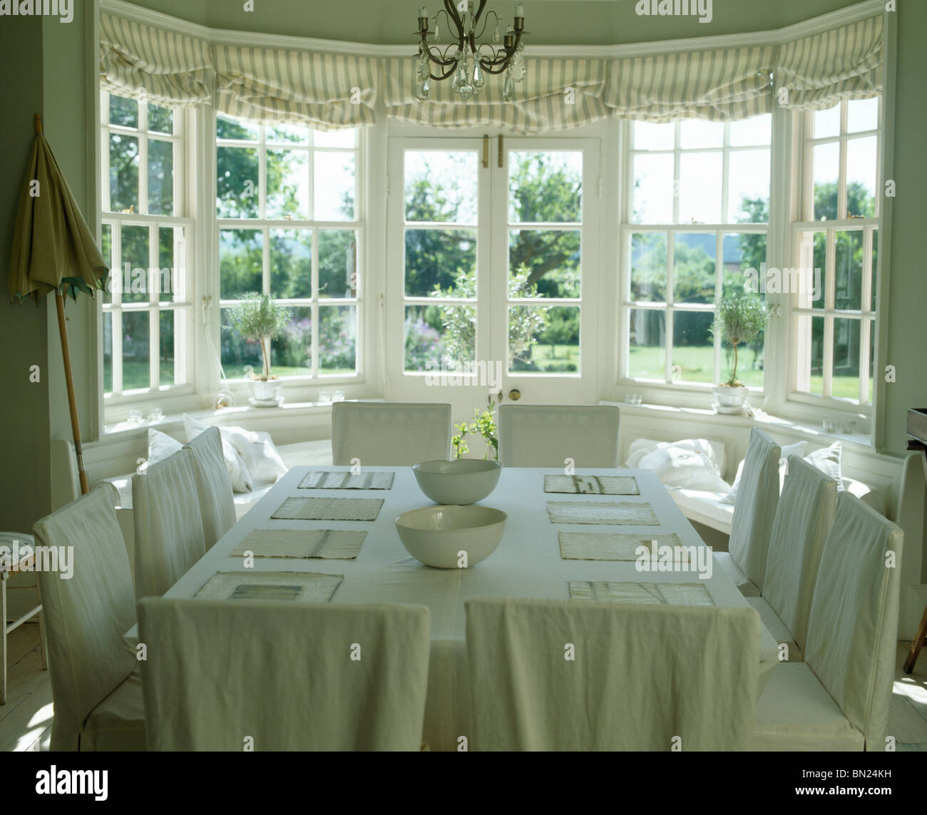 Linge de crème-couvre lâche sur des chaises de salle à manger pays crème avec tissu en lin sur table en baie vitrée avec vue sur le jardin Banque D'Images