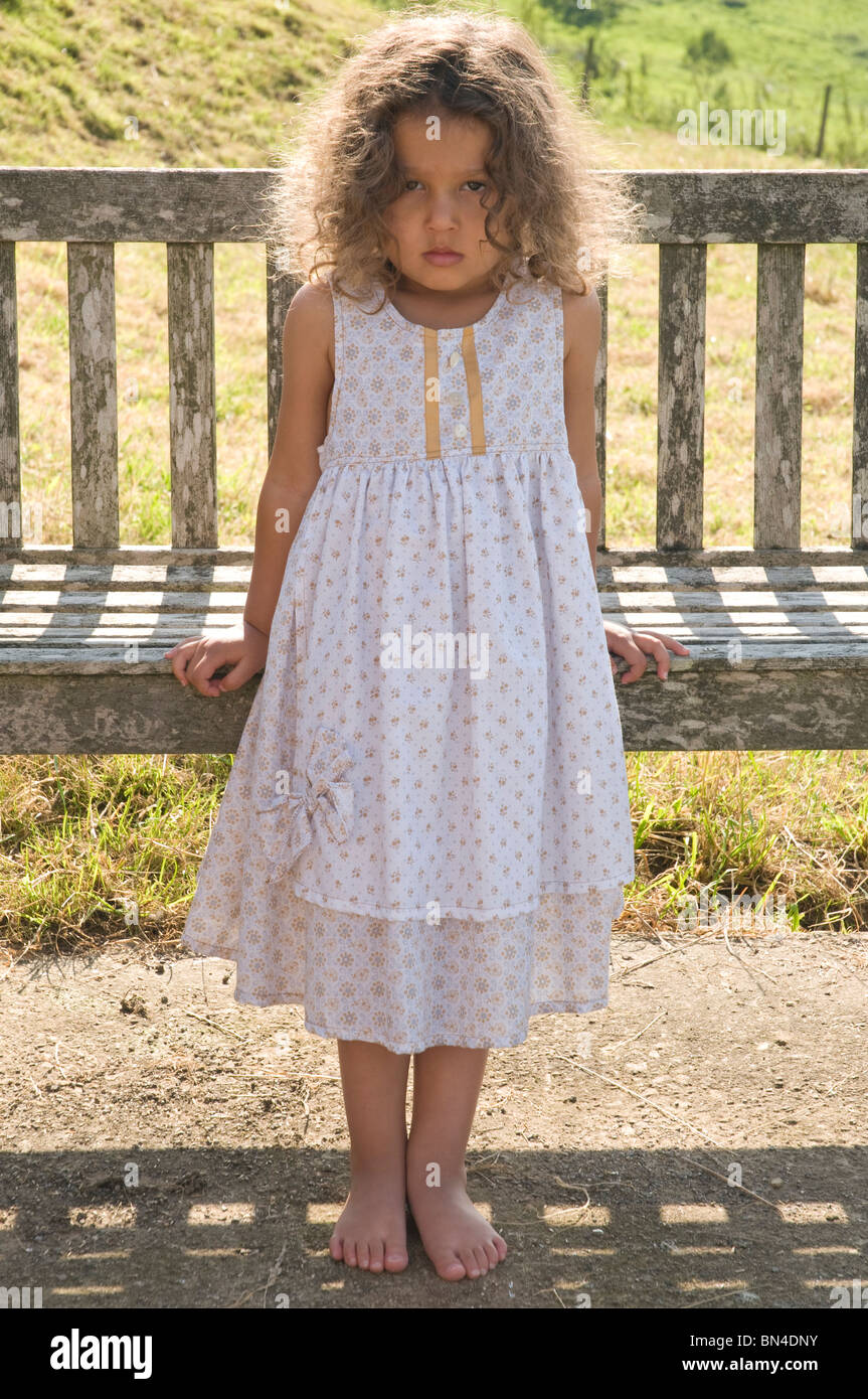 Petite fille aux cheveux bruns weaing une robe d'été appuyé contre un banc en bois Banque D'Images