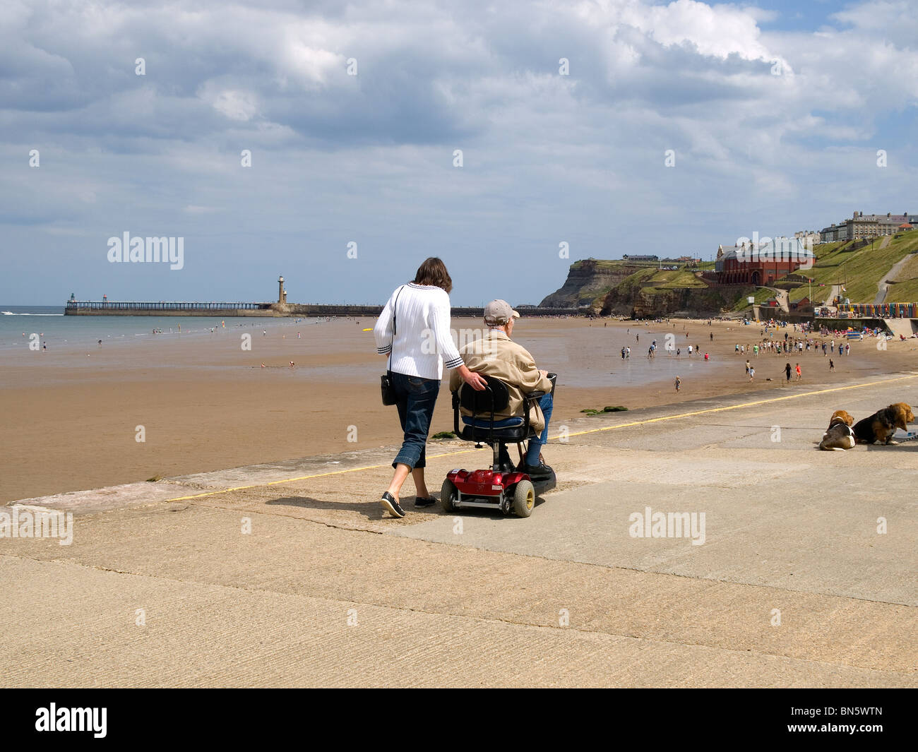 Une jeune femme marche à côté d'un très vieil homme sur un scooter de mobilité à Whitby, North Yorkshire Banque D'Images