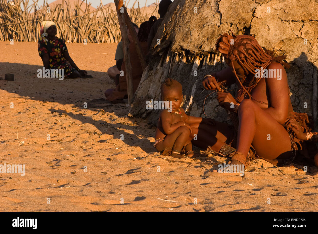 Les nomades de l'Afrique mère Himba et bébé assis dans le sable au coucher du soleil par de la bouse de vache hut travailler avec perles en costume traditionnel du désert de Namibie Banque D'Images