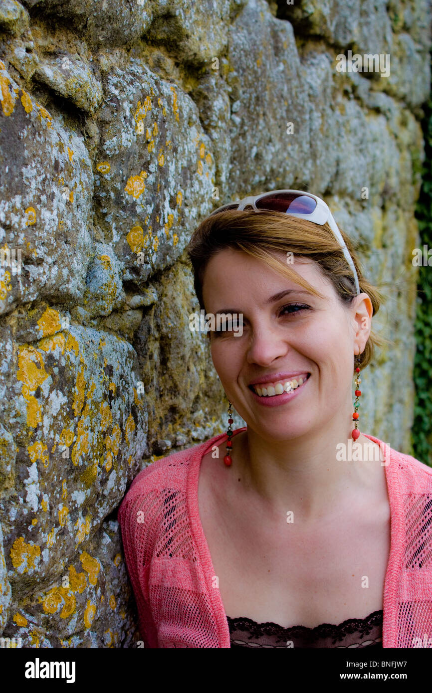 Smiling attractive dame appuyée contre un vieux mur en pierres usées météo. Elle est au début de la trentaine. Banque D'Images