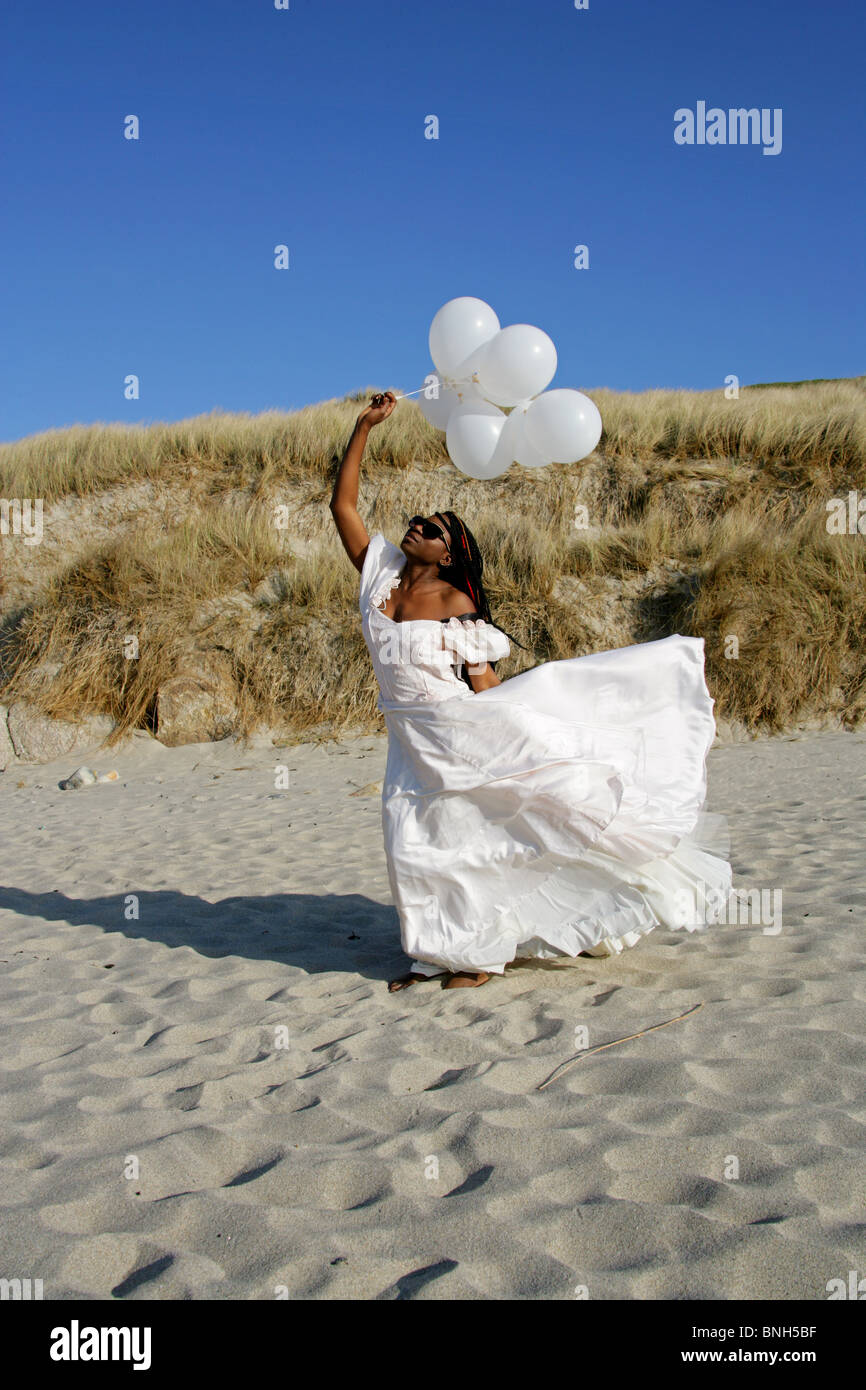 Femme africaine avec des dreadlocks, vêtu d'une robe de mariée blanche, Holding Balloons et debout sur la plage par les dunes de sable. Banque D'Images