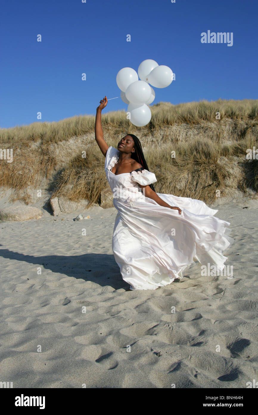 Femme africaine avec des dreadlocks, vêtu d'une robe de mariée blanche, Holding Balloons et debout sur la plage par les dunes de sable. Banque D'Images