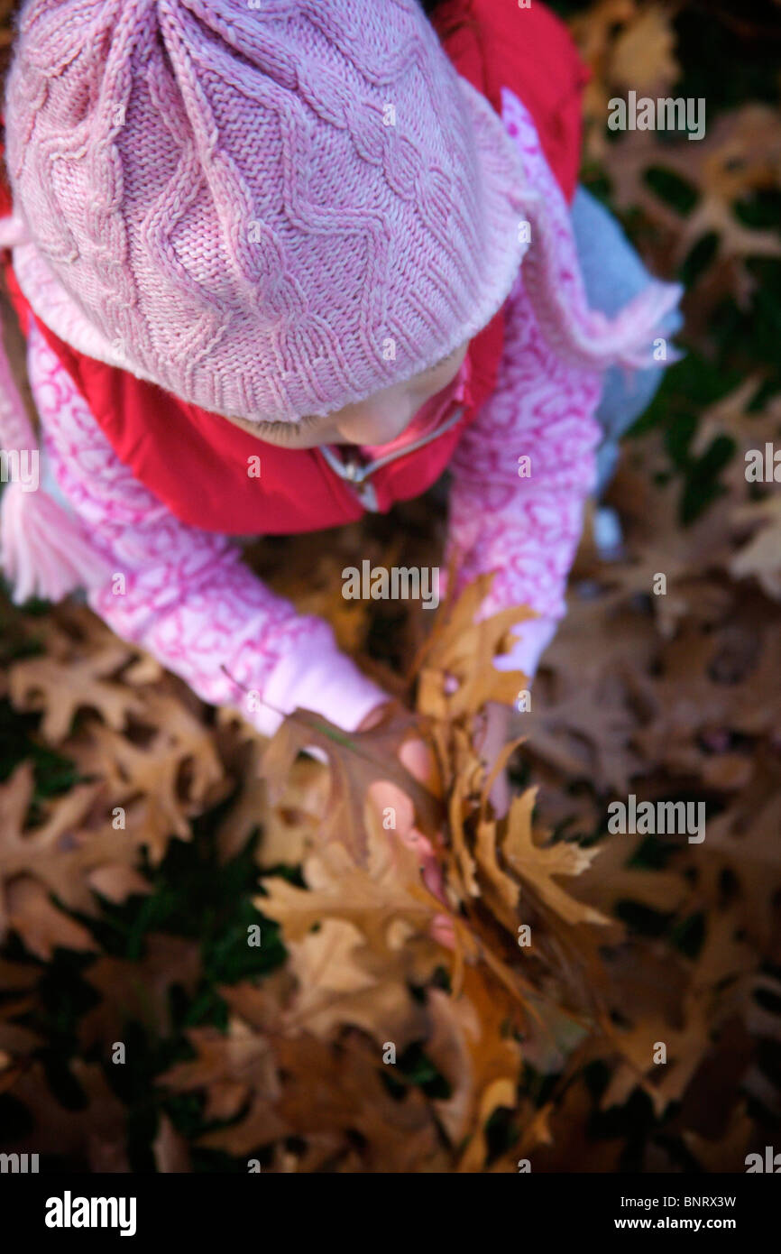 Jeune fille portant bonnet rose et gilet rose joue avec des feuilles sèches dans une cour. Banque D'Images Jeune fille portant bonnet rose et gilet rose joue avec des feuilles sèches dans une cour. Banque D'Images