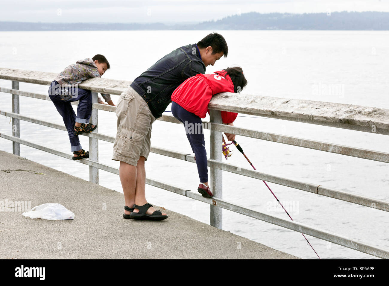 Heureux papa d'Asie montre petite fille comment gérer rod sur la jetée de pêche d'Edmonds sous un ciel gris alors que les jeunes fils watches Banque D'Images