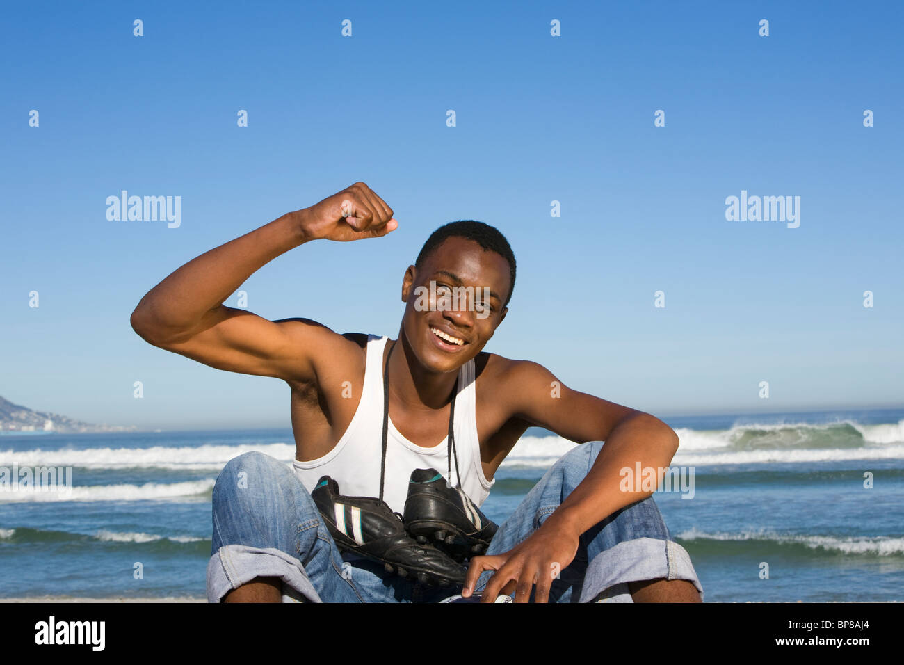 Homme assis sur la plage avec des chaussures de foot autour du cou cheering Banque D'Images