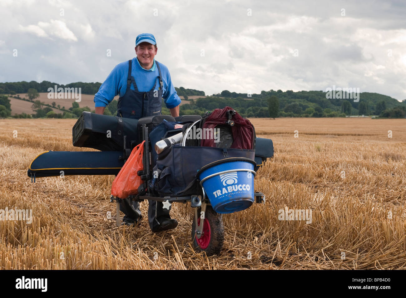 Joyeux pêcheur poussant une brouette de pêche chargée avec du matériel de pêche faisant son chemin vers un champ de maïs chaume vers le bord de la rivière Banque D'Images