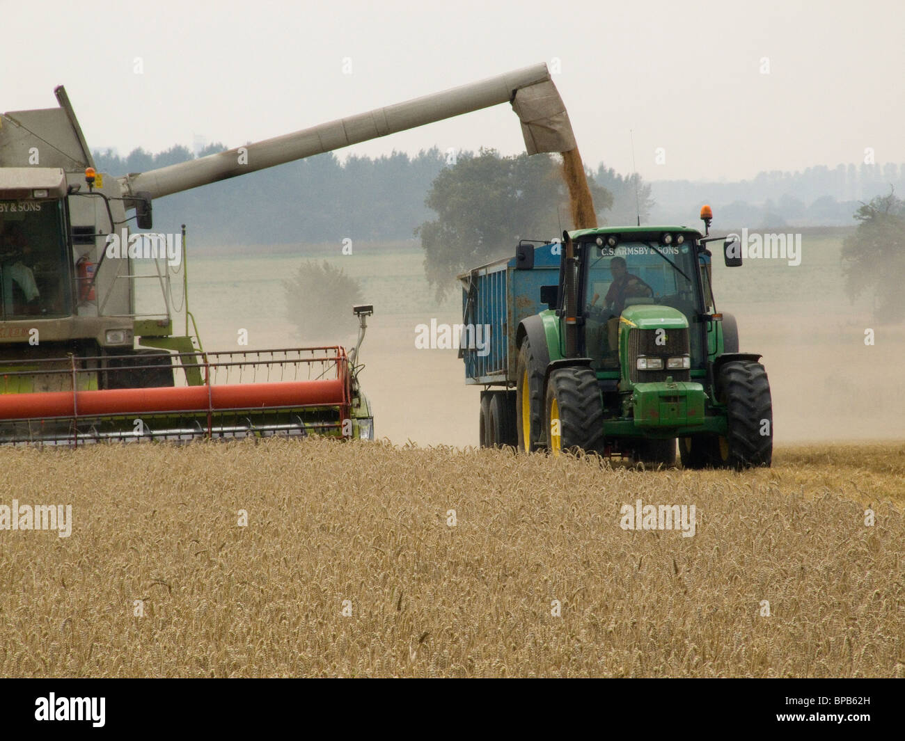 Moissonneuse-batteuse Claas le déchargement du grain dans une remorque remorqué par un tracteur John Deere sur un jour d'été légèrement nuageux Banque D'Images