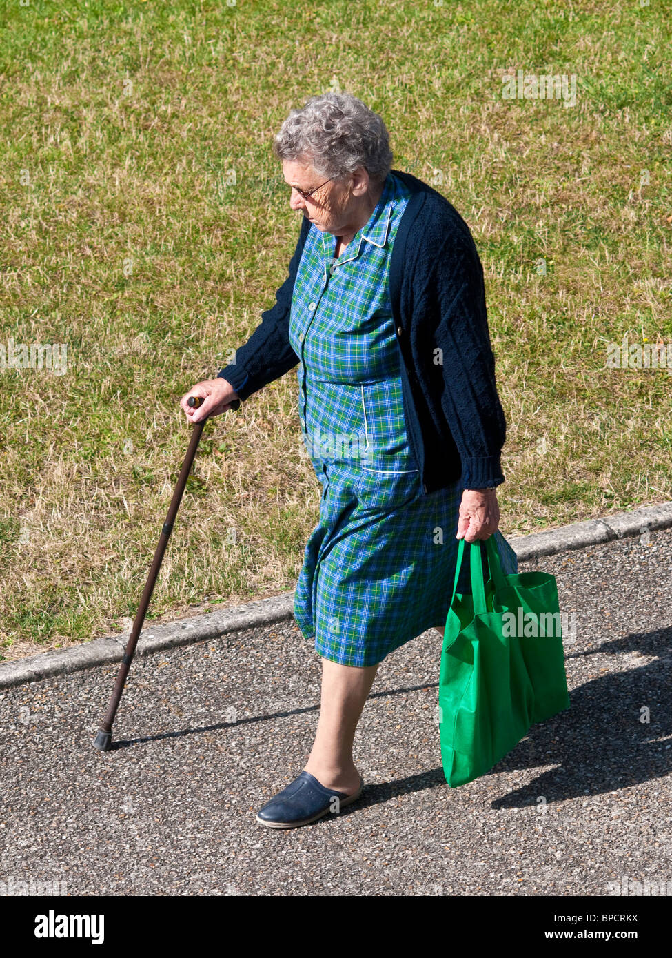 Femme plus âgée avec panier et stick marche le long trottoir - France. Banque D'Images