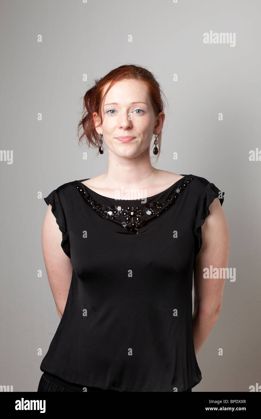 Femme aux cheveux rouges en blouse noire avec encolure en perles souriant doucement tout en se tenant devant un mur clair Banque D'Images