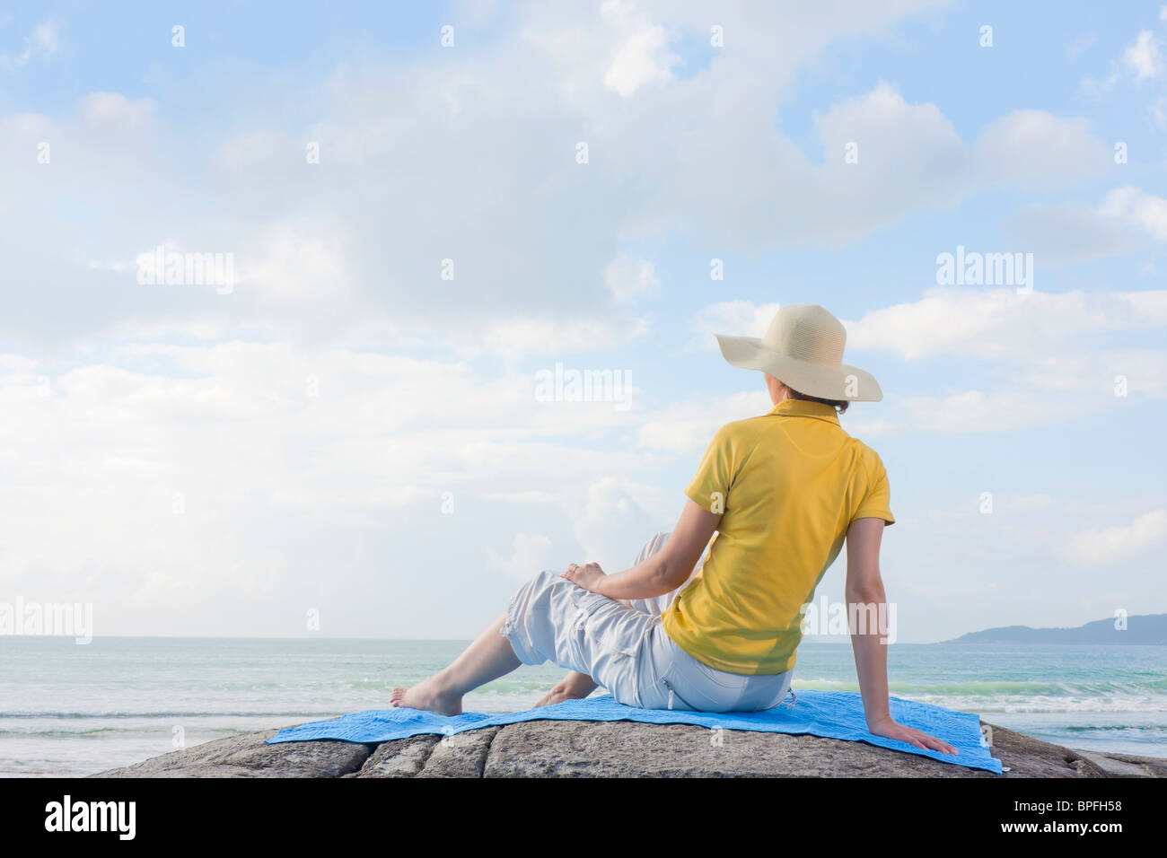 Mid-adult woman with straw hat assis sur un rocher face à la mer Banque D'Images