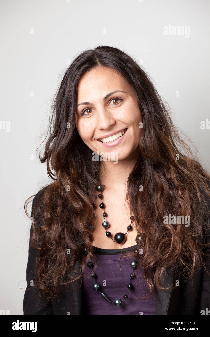 Femme souriante avec de longs cheveux bouclés portant un blazer noir et collier de perles, posant avec confiance dans un plan de studio. Banque D'Images