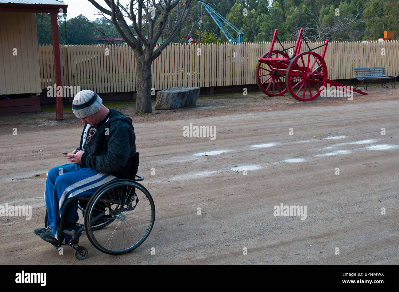 Homme handicapé en fauteuil roulant utilisant un téléphone portable pour envoyer des SMS juxtaposés contre un chariot de chargement de fret antique dans le port fluvial d'Echuca Banque D'Images