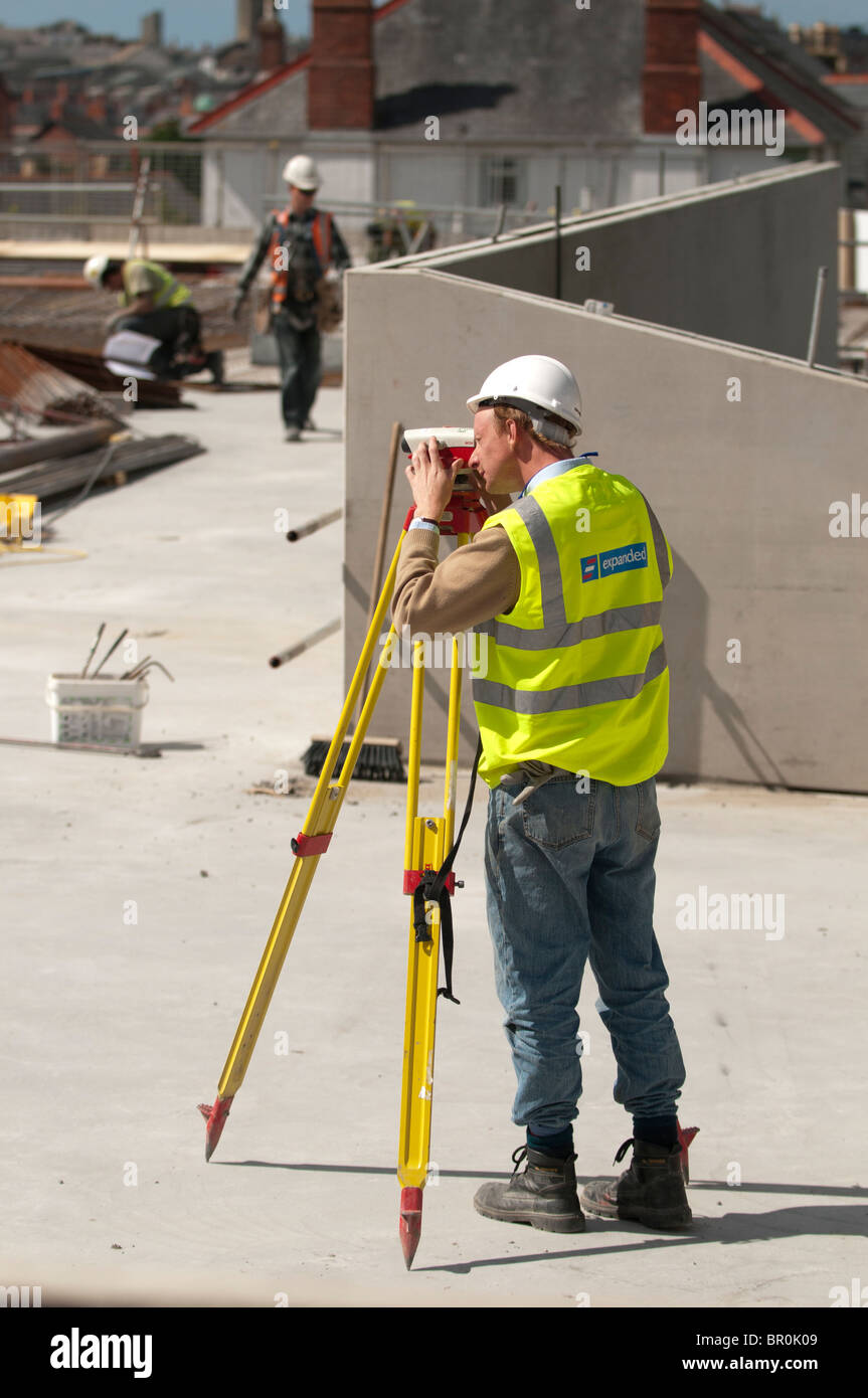 Un homme, vêtu de la Veste haute visibilité et équipement de protection individuelle complète, travaillant sur un chantier de l'arpentage de l'emplacement avec un théodolite Leica, UK Banque D'Images