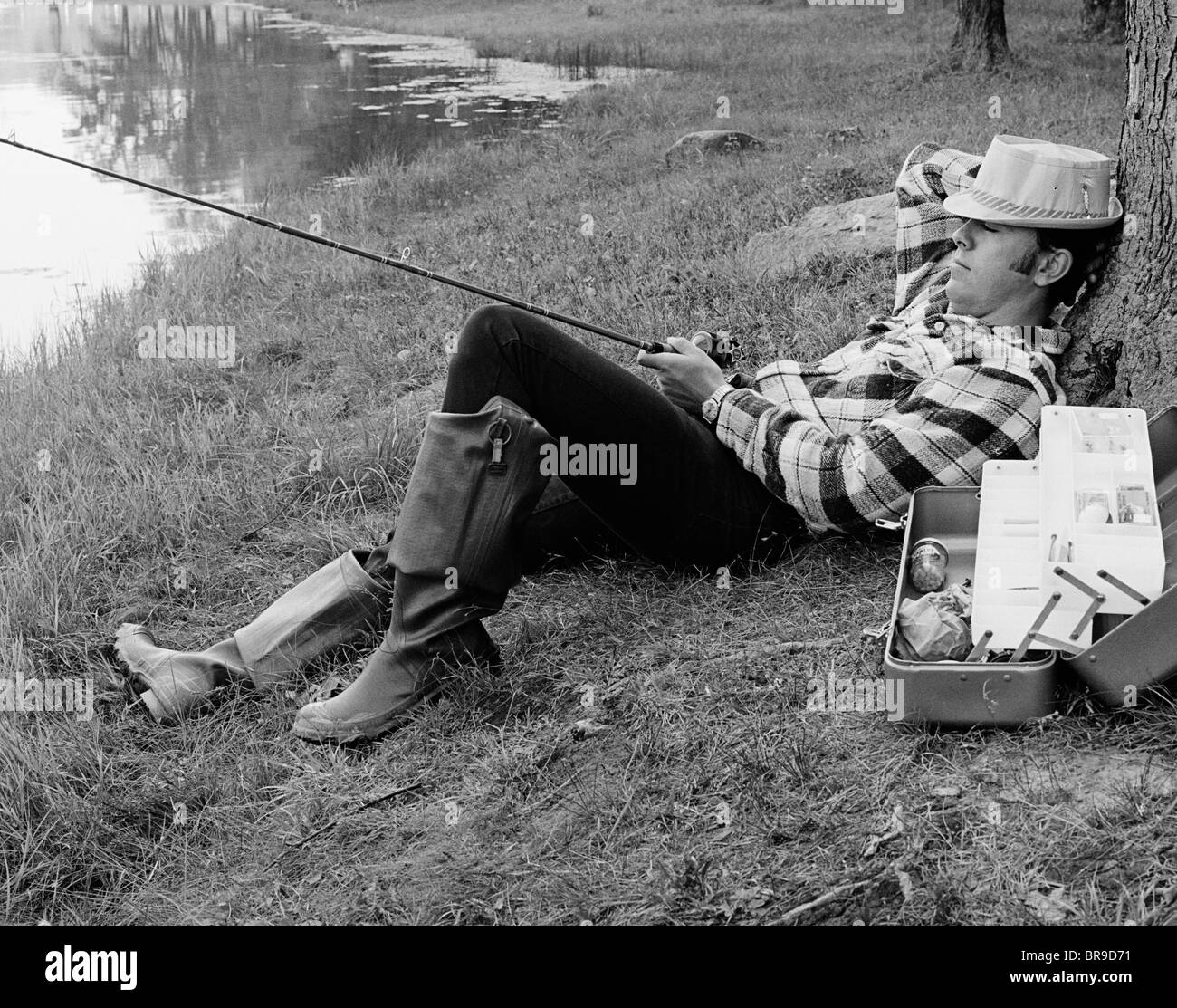 1970 MAN SLEEPING CONTRE UN TRONC D'ARBRE TANDIS QUE LA PÊCHE DANS UN LAC Banque D'Images