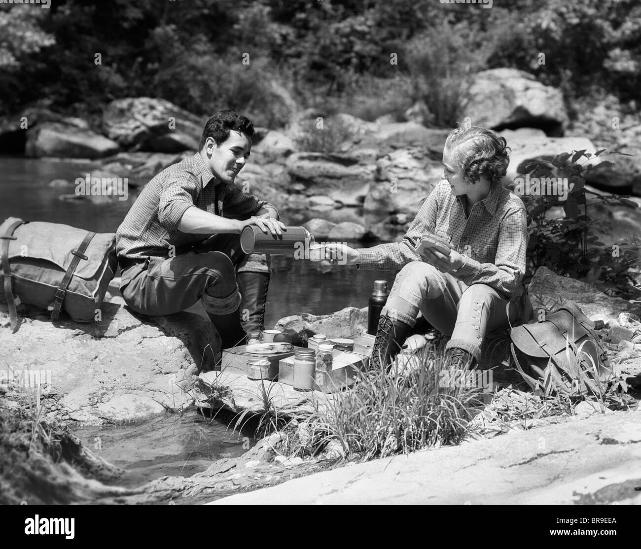 1930 COUPLE HAVING PICNIC ASSIS SUR DES ROCHES PAR STREAM MAN POURING VERRE THERMOS POUR FEMME DE Banque D'Images