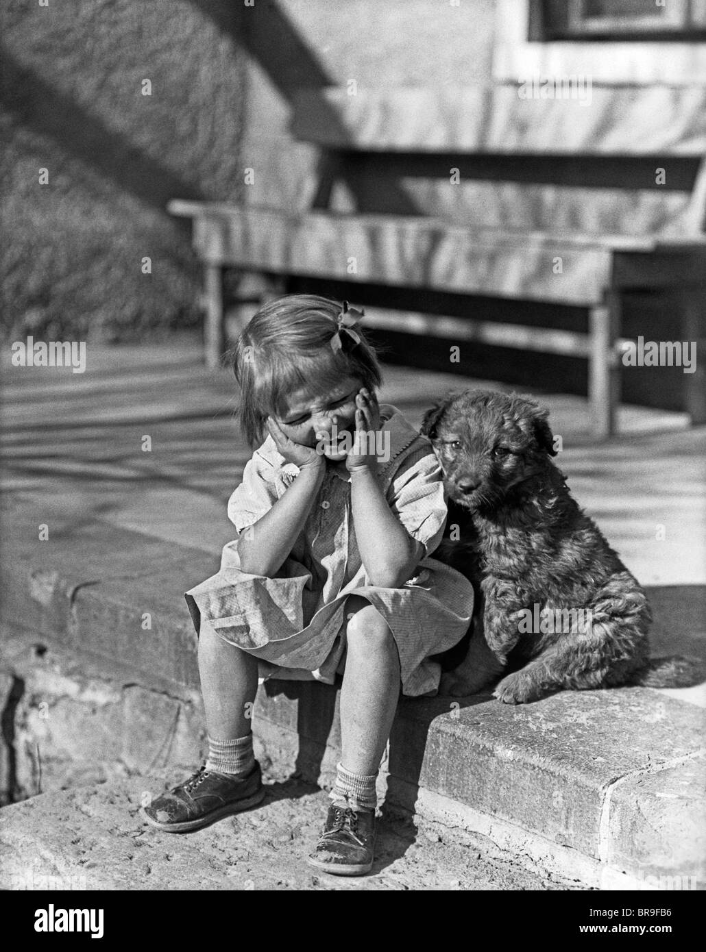 1930 Petite fille assise SUR LE PORCHE STOOP EXPRESSION DROLE MAINS JUSQU'À SON VISAGE À côté de son chien Banque D'Images