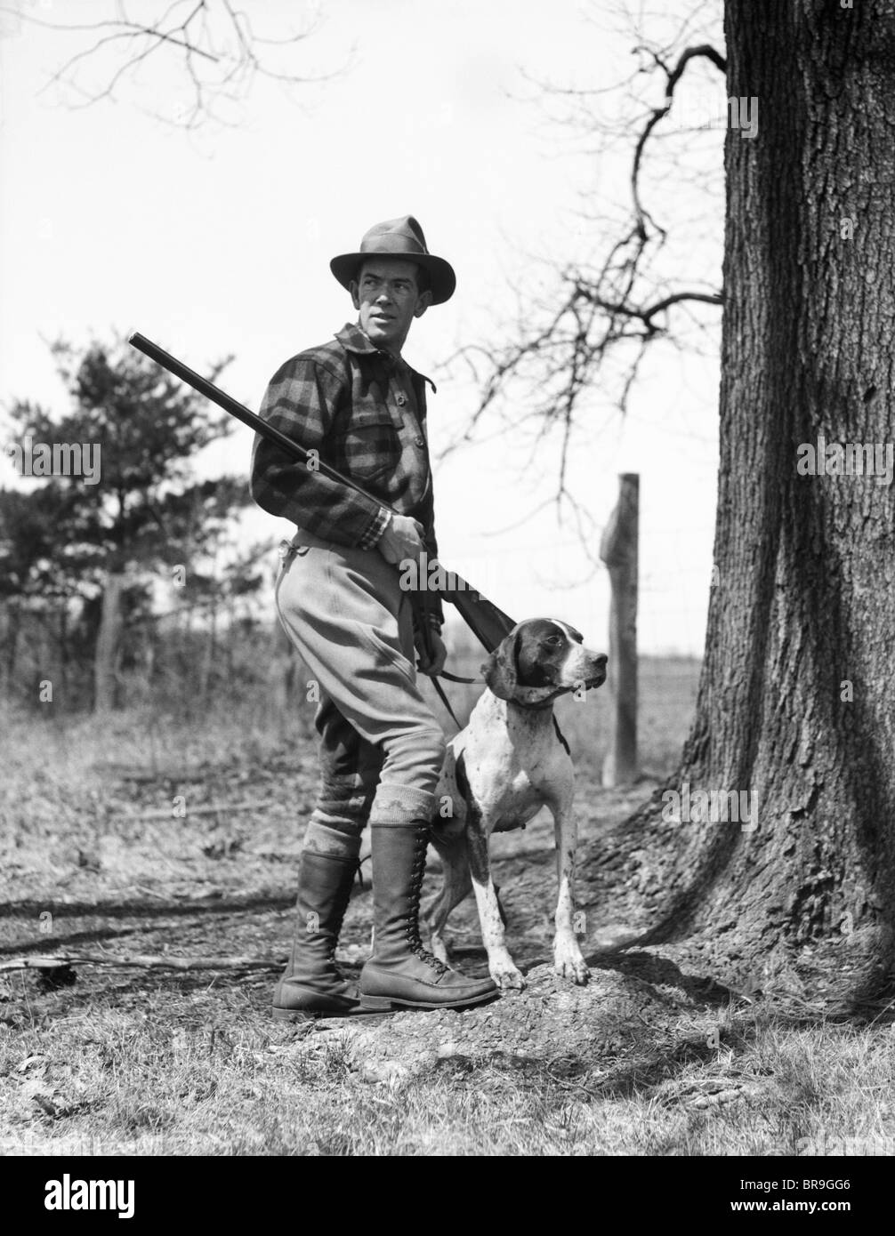 1930 MAN HUNTER WEARING HAT chemise de flanelle à carreaux et des bottes d'équitation fusil de chasse avec l'AIGUILLE DU HOLDING DOG Banque D'Images