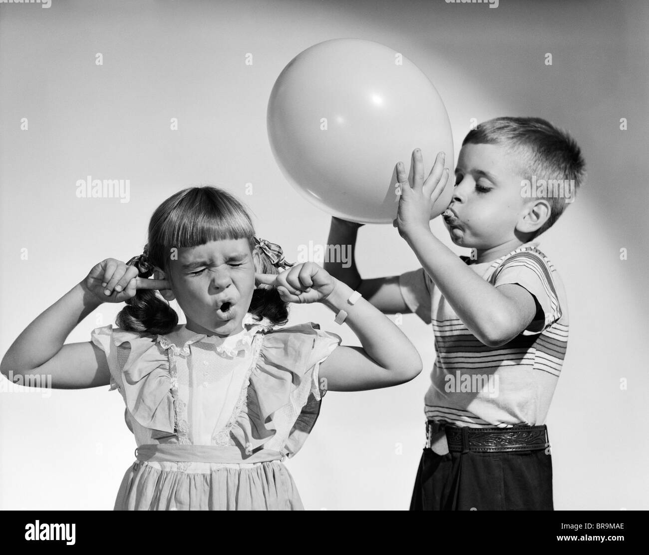 1950 LITTLE BOY BLOWING UP BIG BALLOON PETITE FILLE AVEC LES DOIGTS DANS  LES OREILLES les yeux fermés Photo Stock - Alamy