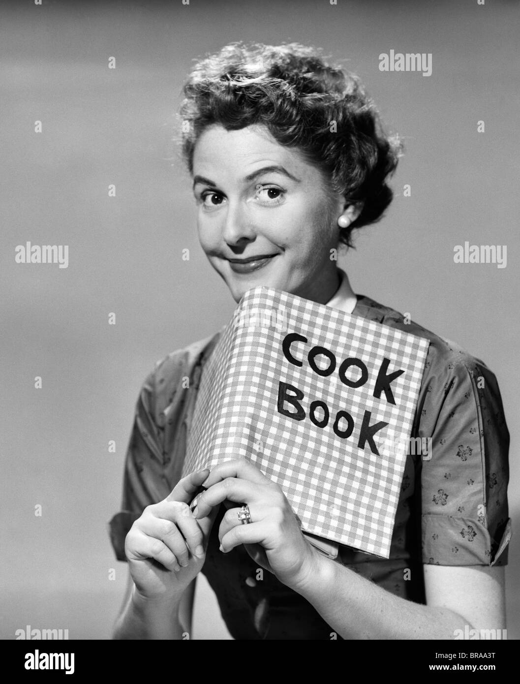 1950 SMILING WOMAN LOOKING AT CAMERA SUR LE DESSUS D'UN LIVRE DE CUISINE Banque D'Images