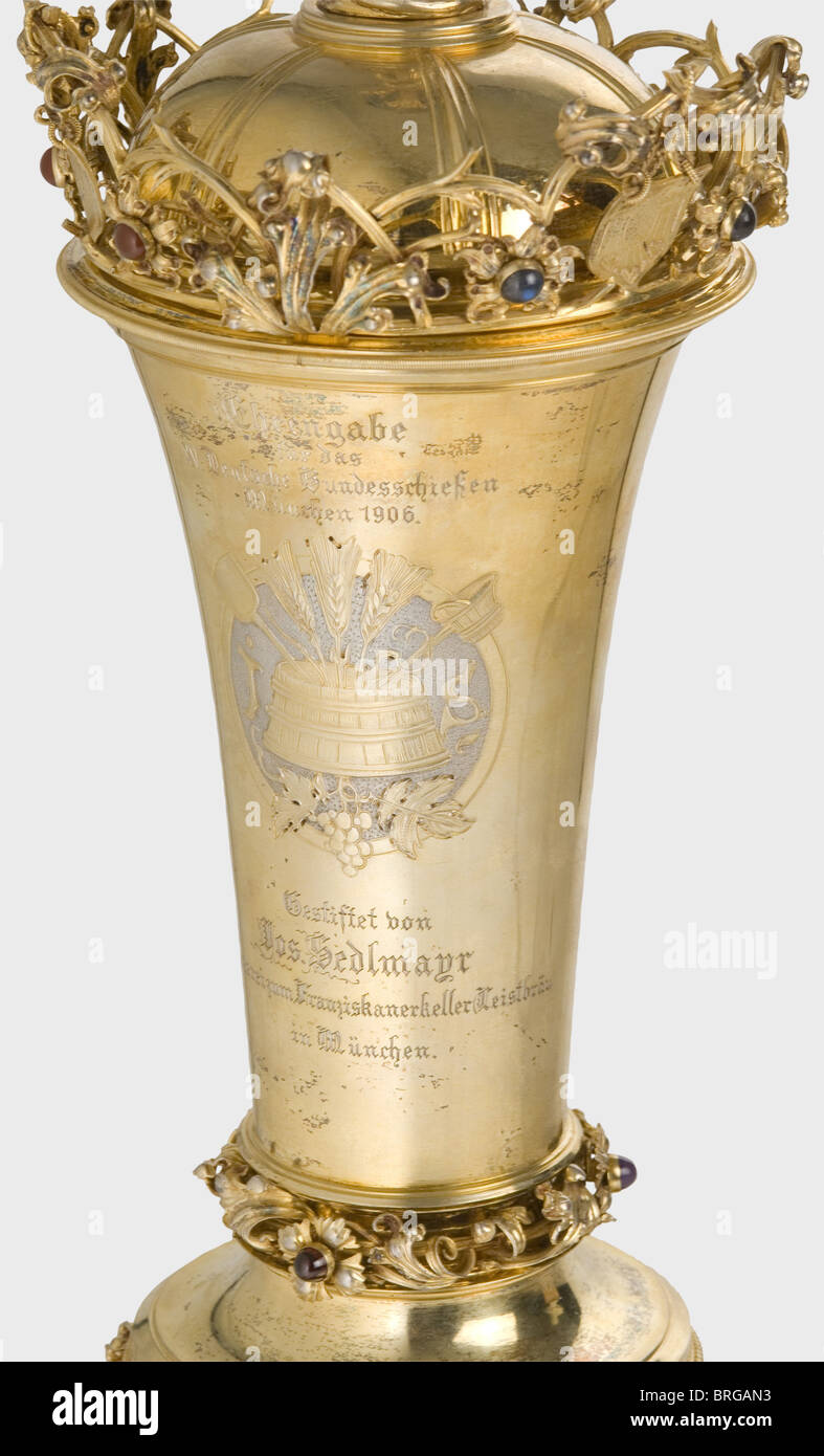 Une coupe trophée, pour le XVème Concours National de tir allemand, Munich 1906 UNE grande, en forme de calice, doré, tasse avec un couvercle.le couvercle porte une boule lapis-lazuli soutenue par des racines avec une miniature argentée de la ville de Munich.le bord est entouré de vigne contenant des boucliers avec des symboles de tir et des fleurs ensemble Sapphires, carnéliens et agates. Le côté opposé de la tasse affiche des outils de brassage entre les initiales 'J' et 'S' et les inscriptions 'Ehrengabe für das XV.Deutsche Bundesschießen München 1906'(Prix pour le XVème Allemand, droits additionnels-Clearences-non disponible Banque D'Images