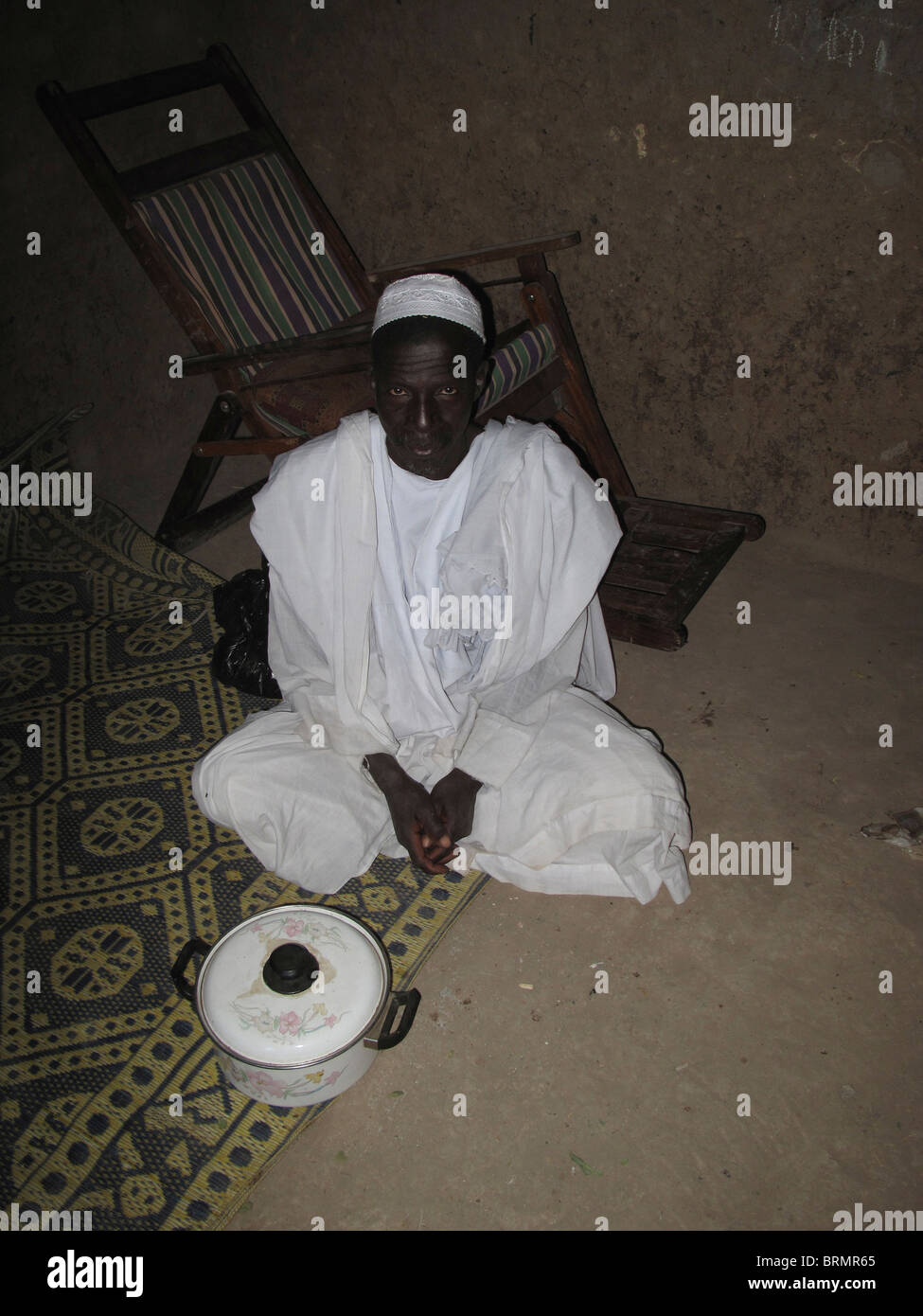 Un adulte homme africain assis sur un tapis avec les jambes croisées portant une robe blanche et calotte traditionnelle Banque D'Images