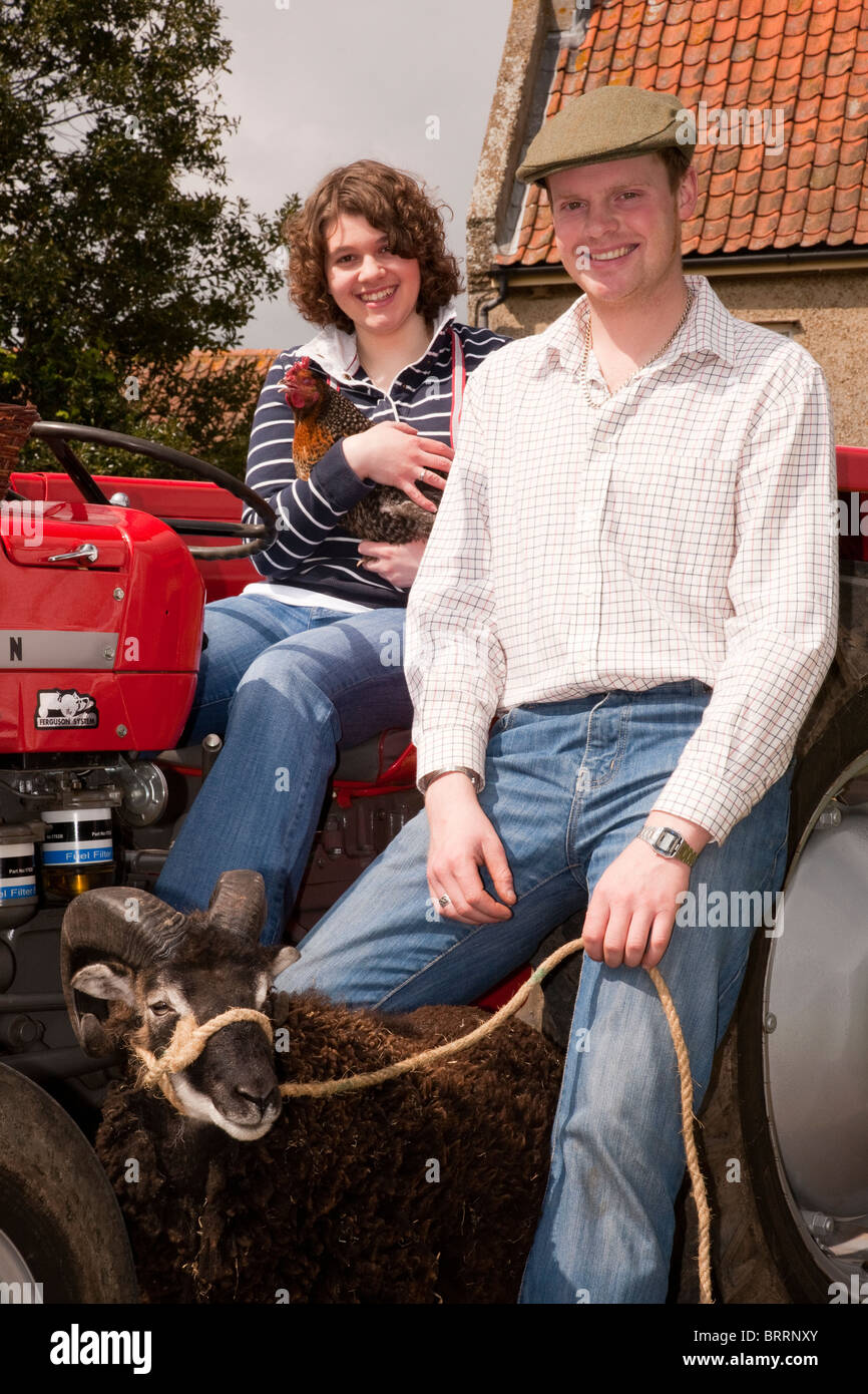 Un couple de jeunes assis sur un tracteur à l'extérieur d'une ferme avec une poule et un bélier Banque D'Images