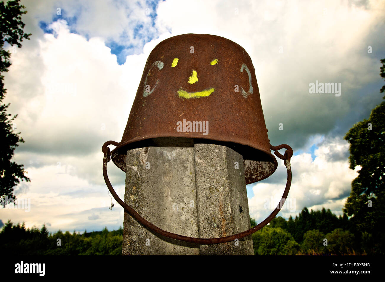 Seau rouillé avec un visage souriant assis au sommet d'un poteau en bois au milieu d'un ciel nuageux Banque D'Images