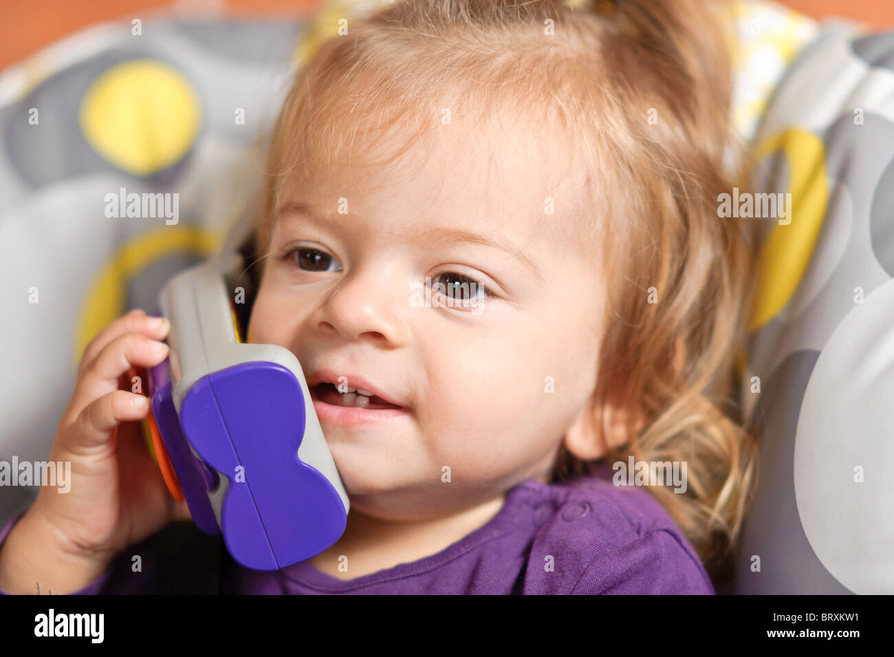 Un bébé fille assis sur une chaise haute fait semblant de tenir une conversation sur un jouet téléphone mobile. Banque D'Images