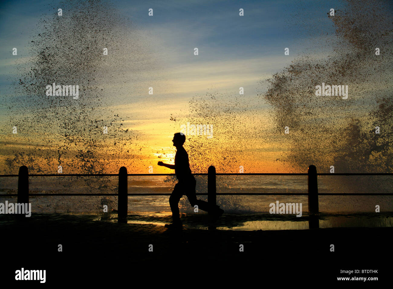 Man jogging sur l'esplanade de Sea Point au coucher du soleil avec des vagues s'écraser sur la côte Banque D'Images