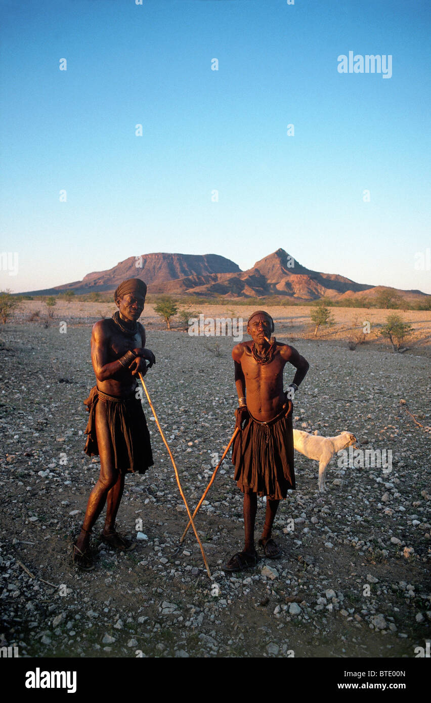 Deux hommes en jupe traditionnelle Himba Standing together appuyée sur de longues cannes de marche avec un chien à l'arrière-plan Banque D'Images