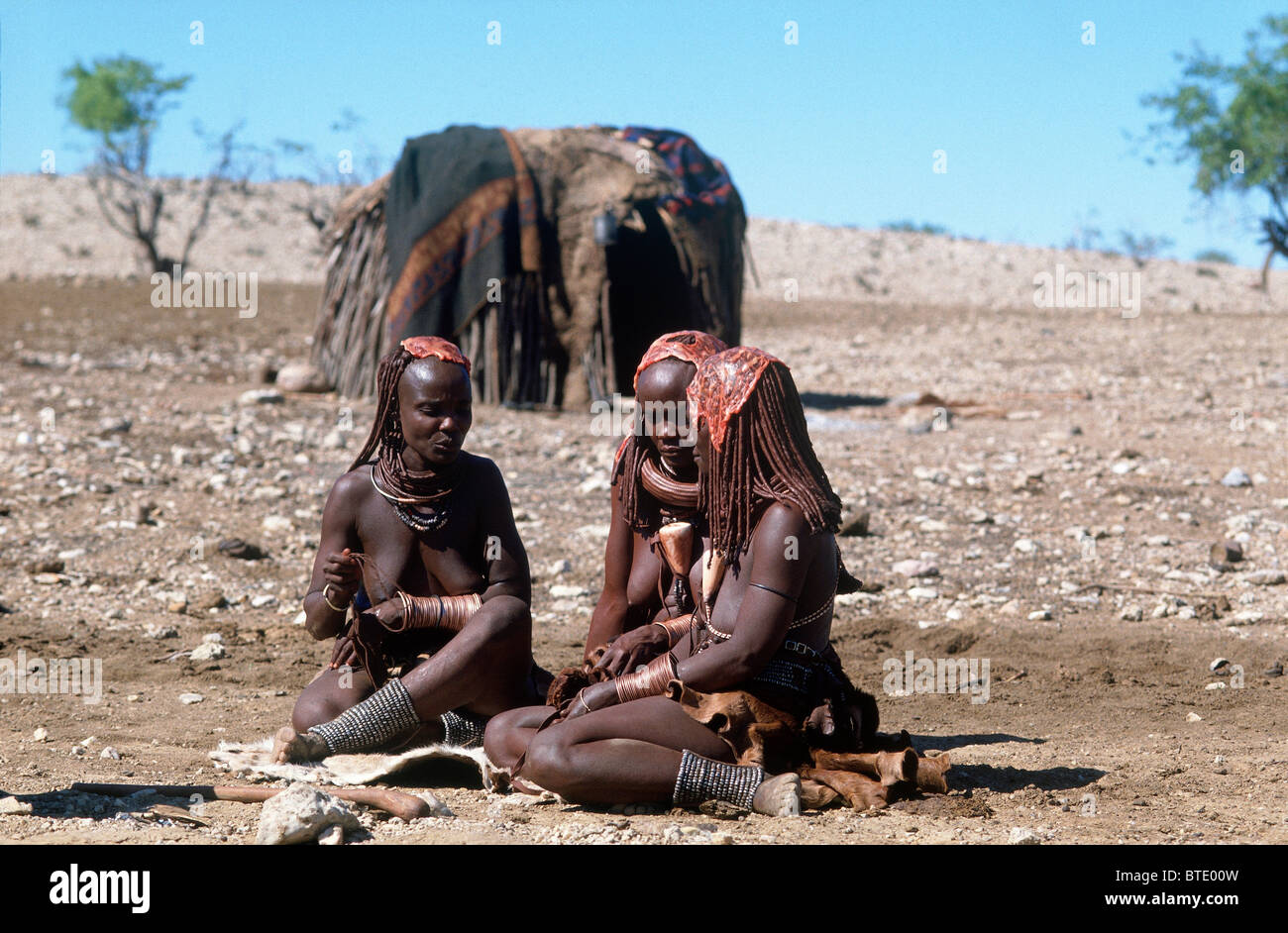 Trois femmes Himba adultes habillés en vêtements traditionnels assis dans un demi-cercle en face de leur bâton et la peau d'animaux accueil Banque D'Images