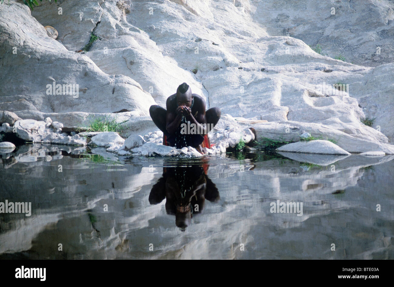 Homme Himba boire d'un flux avec creux des mains et les roches blanches derrière lui Banque D'Images
