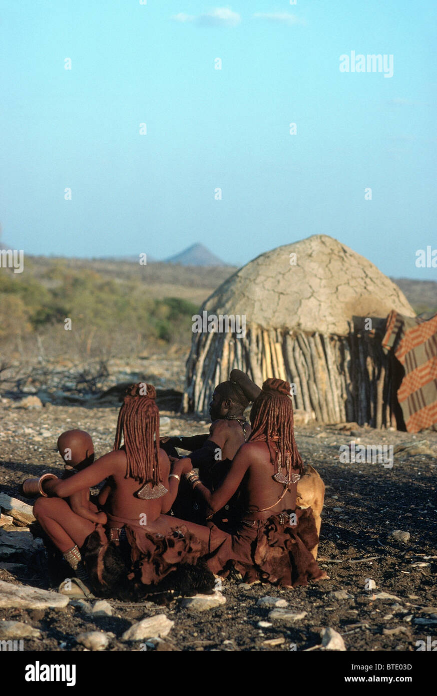 Trois femmes Himba et un jeune enfant assis sur le sol près d'une hutte traditionnelle Banque D'Images