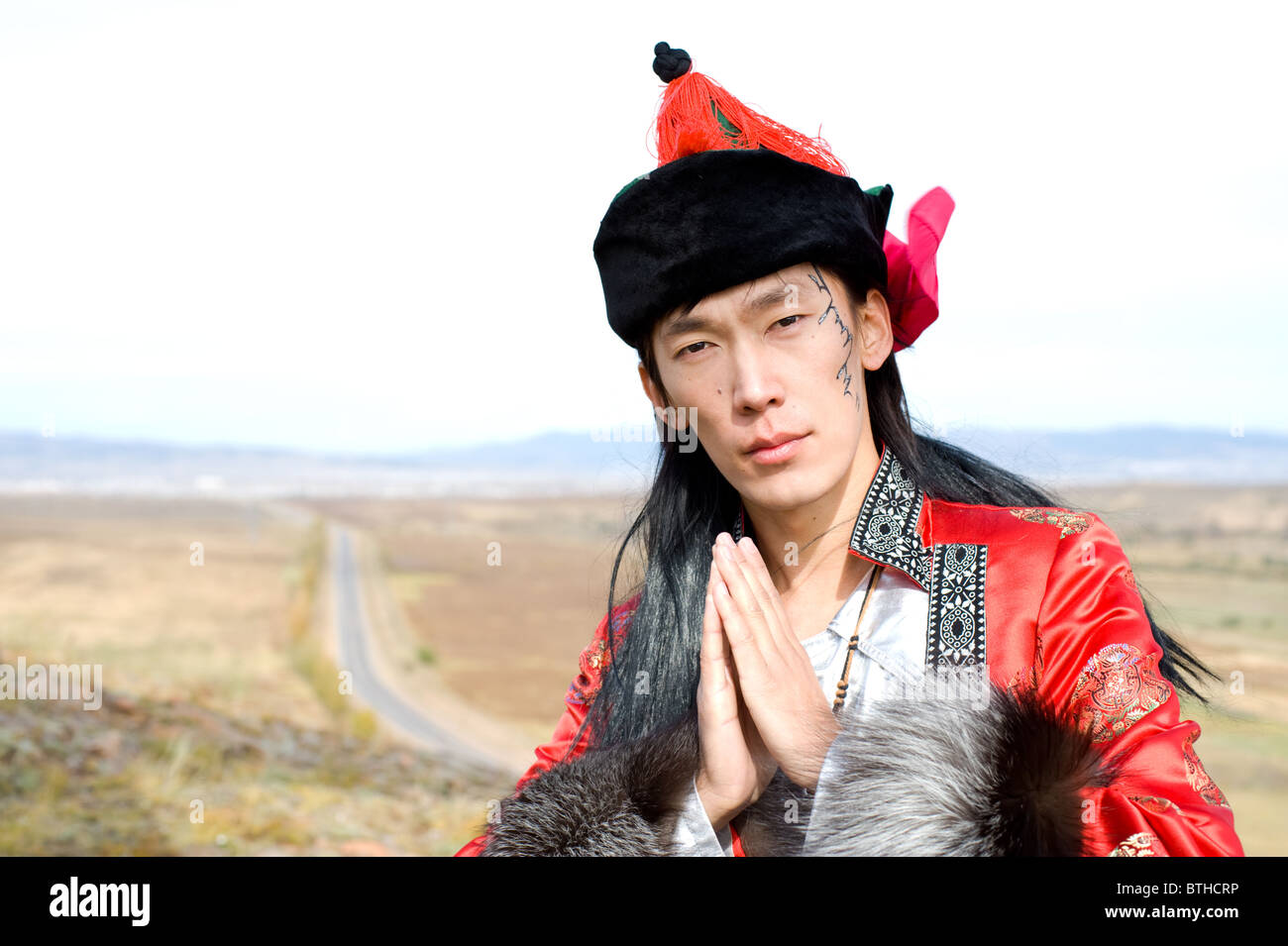 Young Asian man in Mongolian costume avec les mains jointes en prière, son nom Chinghis est peint en mongol ancien sur son visage Banque D'Images