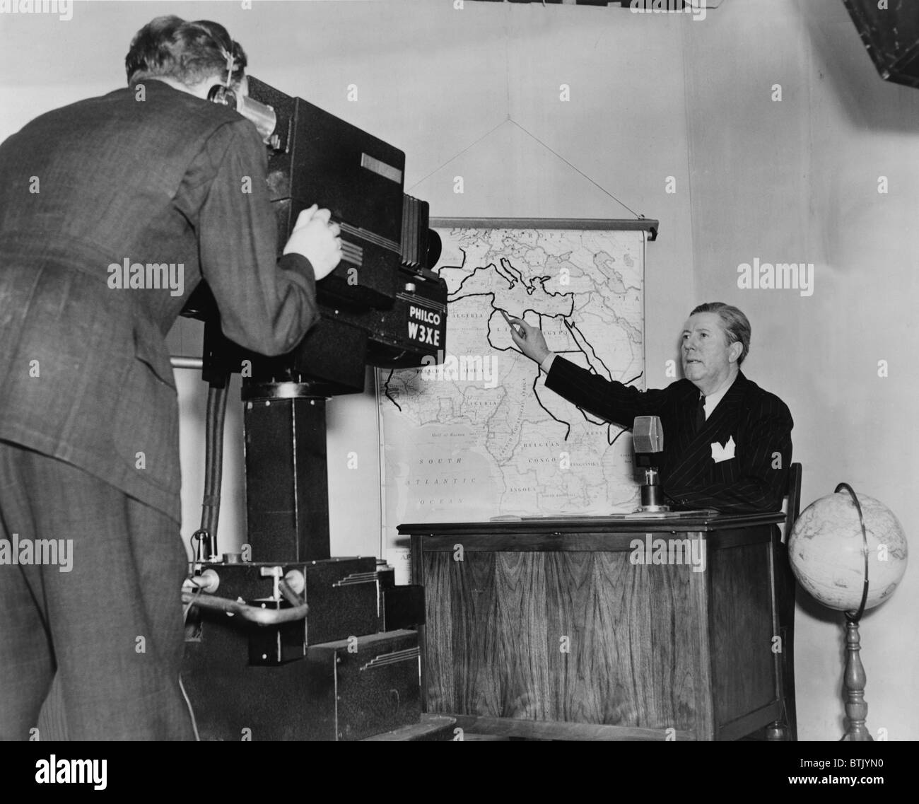 Frazier Hunt, (1885- ) début de journaliste, assis au bureau, en pointant sur une carte murale de l'Afrique, tout en étant filmé par un caméraman. 1941. Banque D'Images
