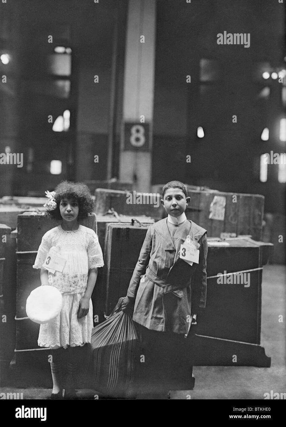 Deux enfants immigrants bien habillé avec le tag 'ERIE' étiquetés à Ellis à l'intérieur des terres, en face d'un grand nombre de lignes de vapeur. Ca. 1910. Banque D'Images