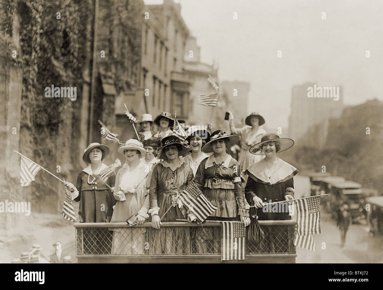 Souriante jeune femme brandissant des drapeaux du haut du véhicule dans un New York City parade. Ca. 1910-1915. Banque D'Images