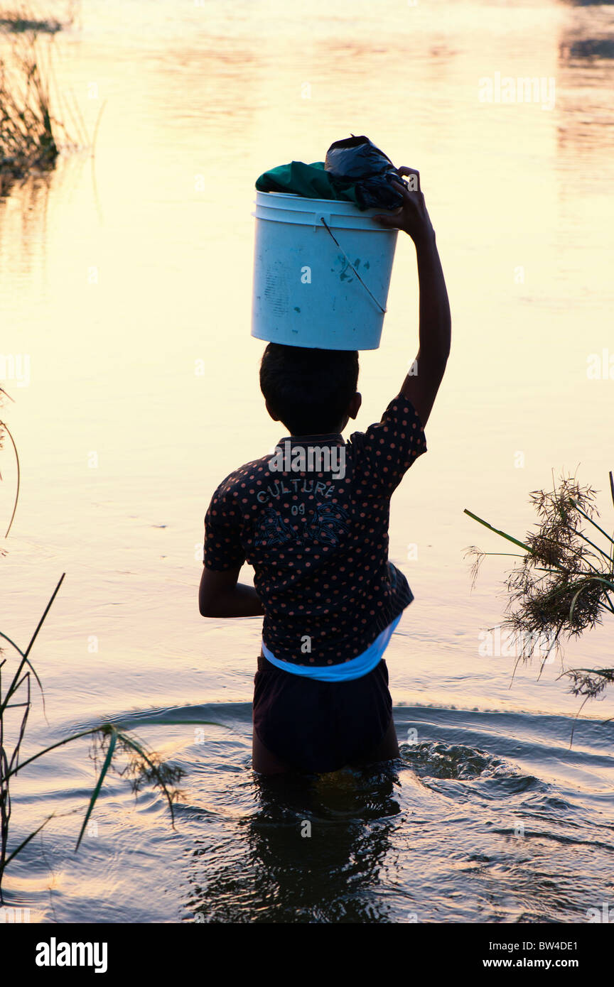 Silhouette d'un garçon portant un seau de lavage sur sa tête à travers une rivière en Inde au lever du soleil. Puttaparthi, Andhra Pradesh, Inde Banque D'Images