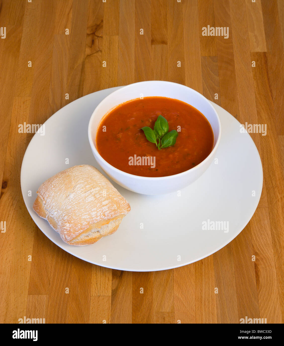 La nourriture, cuit, soupe, bol de soupe tomate et basilic sur une plaque avec un pain rustique de rouler sur une surface de table en bois. Banque D'Images