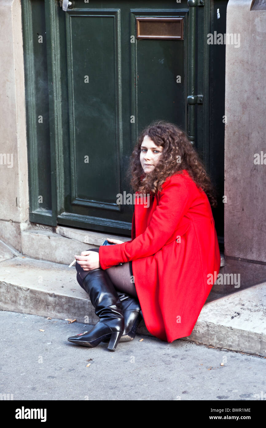 Jolie étudiante aux yeux bleus avec de longs cheveux curling portant veste rouge vif assis sur stoop fume cigarette Rue Saint Jacques Banque D'Images