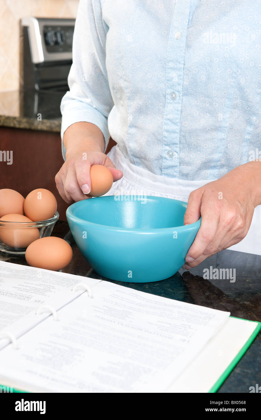 Une femme au foyer se fissure un oeuf dans un bol à mélanger tout en suivant les instructions d'un livre de cuisine. Banque D'Images
