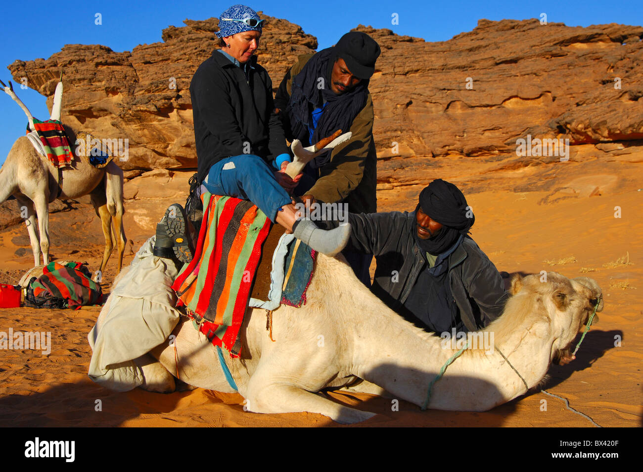 Les nomades touaregs aider un touriste femelle à monter sur un dromadaire dans le désert du Sahara, montagnes Acacous, Libye Banque D'Images
