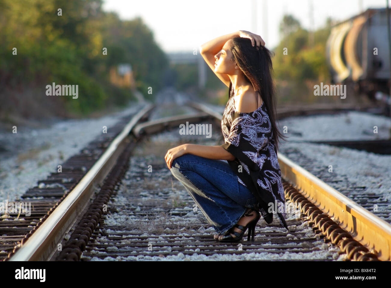 Une belle latina adolescents s'accroupit sur une voie ferrée, face au soleil, les yeux fermés. Banque D'Images