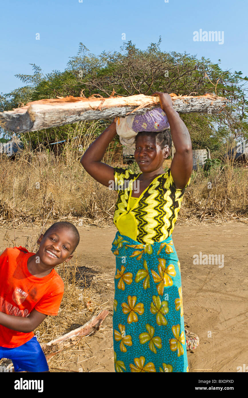 Femme porte le bois pour cuire les incendies dans son village, Kawaza, la Zambie, l'Afrique. Ces femmes sont de la tribu Kunda. Son fils sourit Banque D'Images