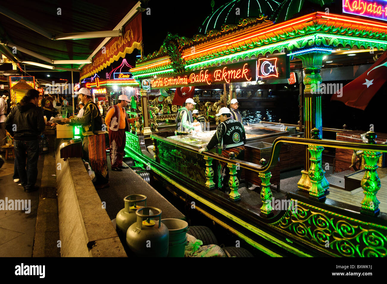 Balik Ekmek restaurants flottants Eminonu Istanbul Turquie // ISTANBUL, Turquie — les bateaux éclairés de balik ekmek servent des sandwichs de poisson traditionnels aux convives le long du front de mer d'Eminonu près du pont de Galata. Ces restaurants flottants, connus localement sous le nom de balıkçı tekneleri, proposent du maquereau grillé servi dans du pain blanc avec des oignons et de la laitue comme nourriture de rue populaire. Les bateaux richement décorés présentent des motifs de style ottoman et restent amarrés en permanence le long du front de mer historique de la Corne d'Or. Eminonu sert de terminal de ferry majeur et de quartier commercial reliant les côtés européen et asiatique Banque D'Images