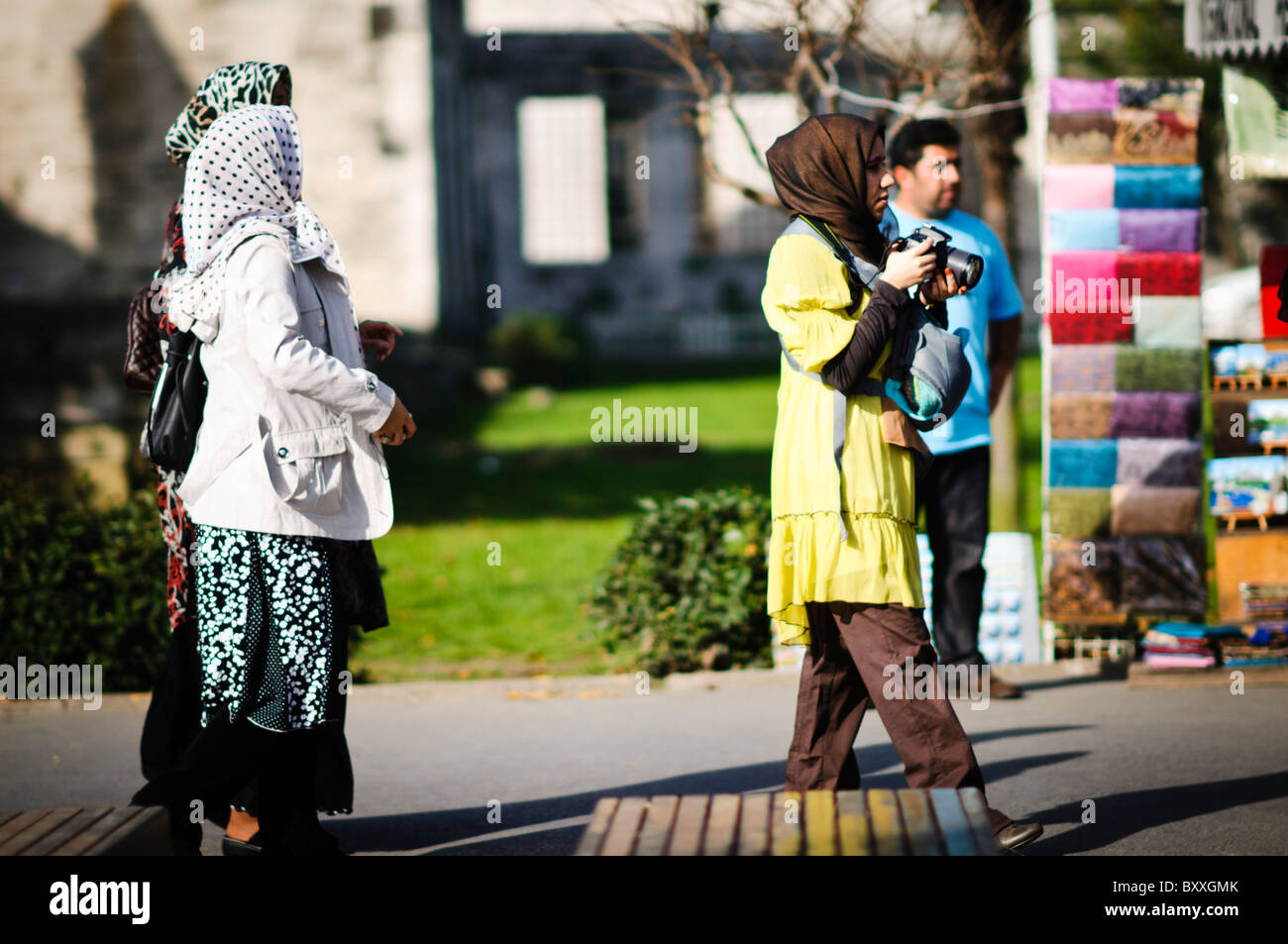 Touristes du moyen-Orient Istanbul Turquie // ISTANBUL, Turquie — des touristes du moyen-Orient explorent la ville d'Istanbul, Turquie. Une femme, portant une robe jaune et un hijab, tient un appareil photo, capturant les vues. D'autres femmes, également en foulard, marchent à proximité, tandis qu'un homme se tient à l'arrière-plan près d'un étal coloré du marché. Banque D'Images