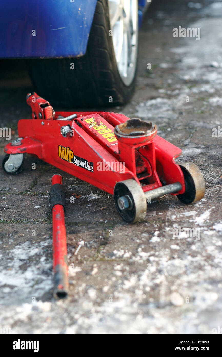 Un gros plan d'un cric rouleur hydraulique rouge à côté d'une voiture sur une allée glacée avec la poignée détachée. Banque D'Images