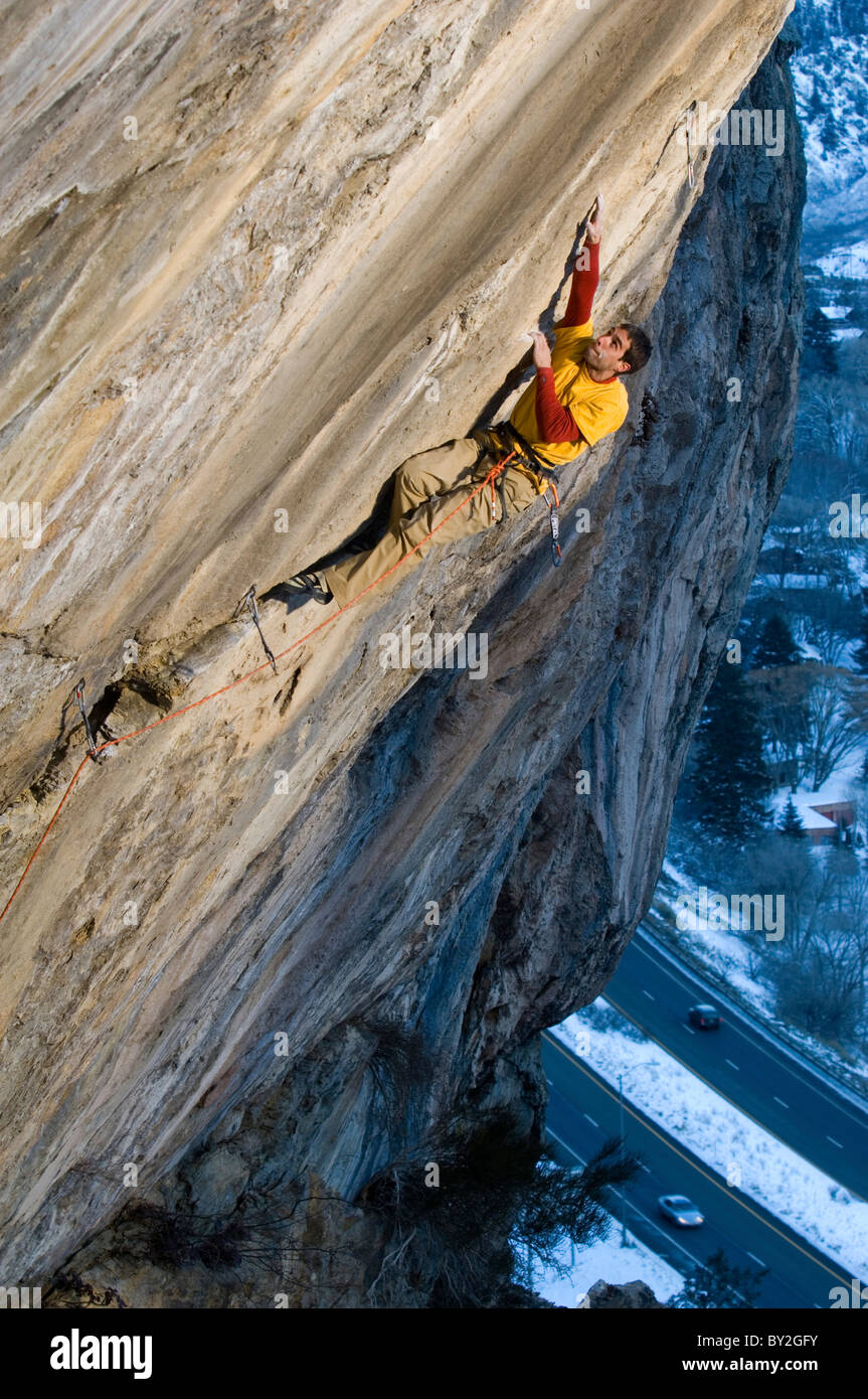 Un alpiniste dans une chemise rouge et jaune atteignant jusqu'à une prise sur une route escarpée et difficile. Banque D'Images