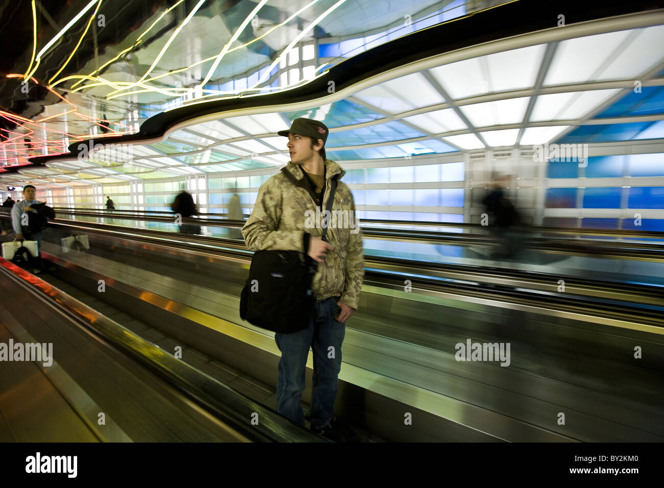 Un jeune homme portant un sac à bandoulière à travers un aéroport sur un système de transfert, comme les néons colorés zigzaguent autour de lui. Banque D'Images